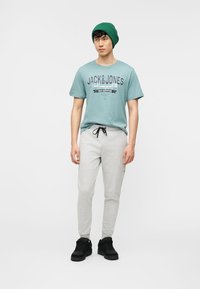 Ung mann iført grønn lue, blågrønn "Jack & Jones" t-skjorte, lysegrå joggebukser med snøring, og svarte sneakers, stående mot hvit bakgrunn.