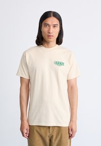 Camiseta de algodón beige con el logo 'OBEY' impreso en verde en el lado izquierdo del pecho. Manga corta, cuello redondo, ajuste relajado y textura suave.