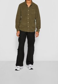 Persona che indossa una camicia a bottoni in corda verde oliva, pantaloni cargo neri e sneakers bianche, in piedi contro uno sfondo grigio chiaro uniforme.