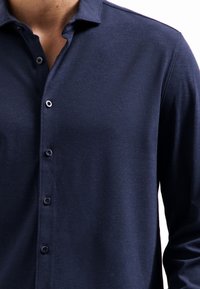 Navyblauw, langemouwhemd van een zachte stof met een spitse kraag. Heeft een knoopsluiting en subtiele textuur.
