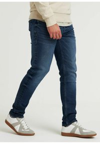 Jeans in denim blu scuro con una vestibilità slim, caratterizzati da tasche imbottite e una trama liscia. Abbinati a sneaker bianche e grigie.
