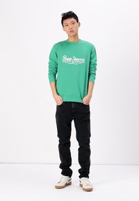 Sudadera verde con logo "Pepe Jeans", de manga larga y cuello redondo; combinada con jeans negros y zapatillas blancas. Diseño simple y casual.