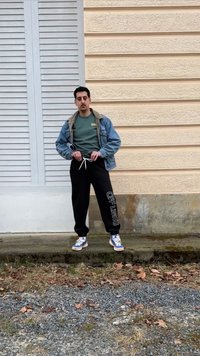 Άνδρας που στέκεται έξω φορώντας τζιν μπουφάν, πράσινο φούτερ, μαύρη φόρμα με το κείμενο "OFF LIMITS" και πολύχρωμα αθλητικά, μπροστά σε μπεζ τοίχο.