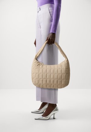 Kobieta w lawendowej bluzce i jasnych spodniach, trzymająca beżową pikowaną torebkę, nosząca srebrne szpiczaste wysokie obcasy.