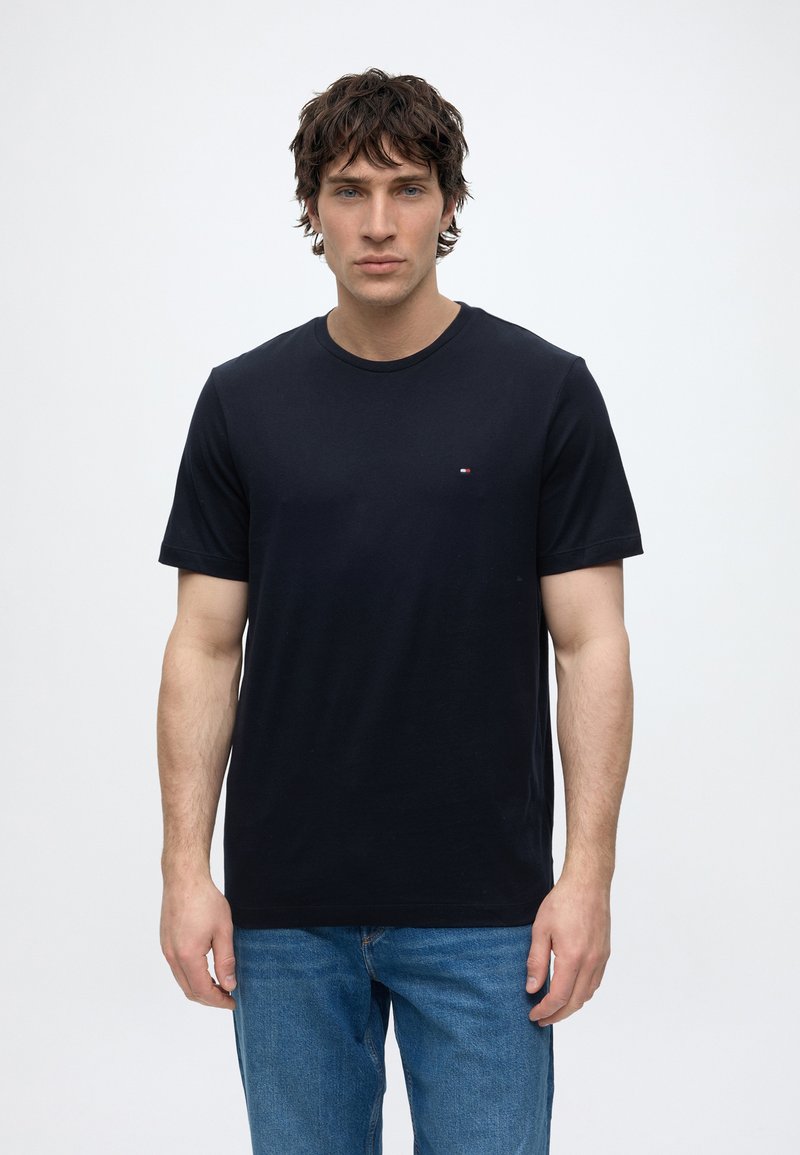 Jeune homme aux cheveux bruns portant un t-shirt noir uni à manches courtes et un jean bleu sur fond blanc.