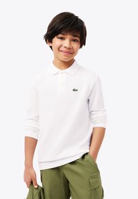 Lächelnder Junge in einem weißen Langarm-Poloshirt mit einem kleinen grünen Logo und olivgrünen Cargo-Hosen, der eine passende Jacke hält.