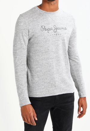 Mężczyzna w jasnoszarej koszuli z długim rękawem z napisem „Pepe Jeans London”, w czarnych spodniach na białym tle.