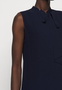 Robe bleu marine sans manches avec un col festonné et un détail noué, en tissu côtelé texturé et coupe ajustée.