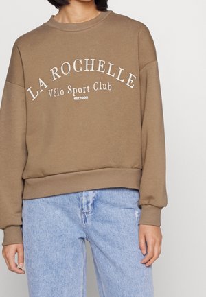 Person trägt einen braunen Pullover mit dem Aufdruck "LA ROCHELLE Vélo Sport Club est. 1998" und hellblaue Jeans vor einem schlichten Hintergrund.