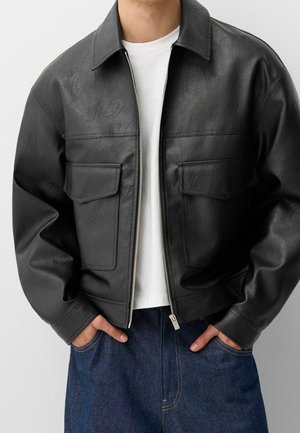 Homme portant une veste en cuir noire ouverte sur un t-shirt blanc avec les mains dans les poches d'un jean bleu.