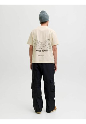Beez värvi puuvillane t-särk mäetipude graafikaga seljal ja "JACK & JONES" tekstiga; koos mustade cargo-pükstega ja pruuni jalatsiga.