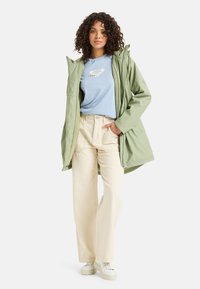 Svetlo zelená nepremokavá bunda s kapucňou, spárovaná s modrým tričkom s logom a krémovo bielymi širokými nohavicami, nosená s bielymi teniskami.