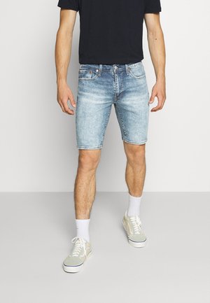 Pantalones cortos de mezclilla azul claro con corte recto, cinco bolsillos y un ligero desvanecimiento. Combinados con calcetines blancos y zapatillas de color claro.