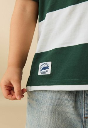 Child's hand beside green and white striped shirt with "Yacht Club & Sailing Goods" label, paired with light blue denim shorts.