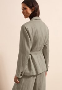 Lichtgrijze blazer van een textuurrijke, vloeiende stof; heeft een ceintuur in de taille, een ingeknipte kraag en knoopmanchetten. Zicht vanuit de achterkant.