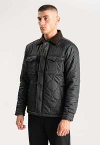 Sort kviltet jakke med knaplukning, der har to brystlommer og en brun fløjlskrave. Glat tekstur og struktureret design.