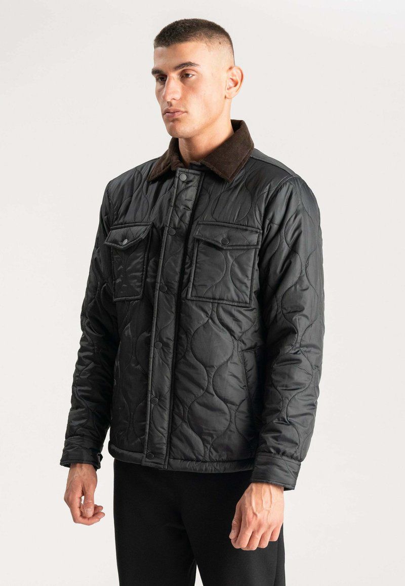 Sort kviltet jakke med knaplukning, der har to brystlommer og en brun fløjlskrave. Glat tekstur og struktureret design.