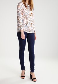 Langärmlige Bluse mit einem floralen Druck in Weiß, Blau und Orange, kombiniert mit dunkelblauen Skinny Jeans und schwarzen Schuhen mit offenen Zehen.