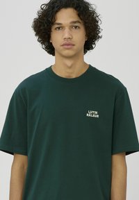 Jeune personne aux cheveux bouclés portant un t-shirt vert foncé avec un petit texte blanc « LUTIN RÂLEUR » sur la poitrine, sur un fond uni.