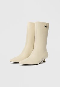 Bottines beiges arrivant au genou faites d'un matériau lisse semblable au cuir, avec un bout pointu, un petit talon et un accent géométrique sur le côté.