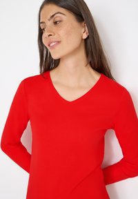 Top rosso a maniche lunghe con scollatura a V. Realizzato in tessuto liscio e elastico. Presenta una silhouette aderente e un semplice dettaglio di cucitura.