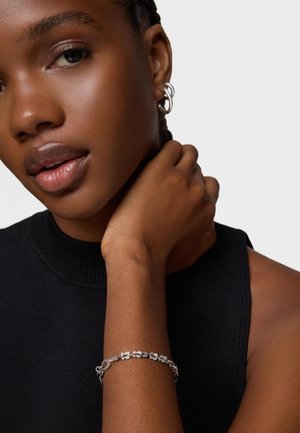 Woman with dark skin wearing silver hoop earrings and a silver floral bracelet, dressed in a sleeveless black top against a light background.