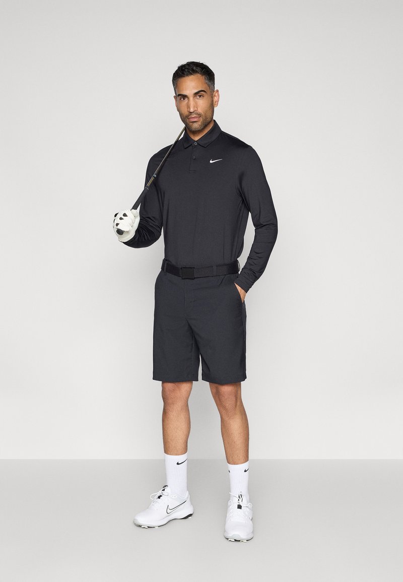 Schwarzes Langarm-Poloshirt mit Kragen, schwarze Shorts, weiße Golfhandschuhe und weiße Schuhe. Nike-Logo auf Shirt und Socken.