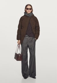 Brun dunjakke med hætte, grå sweater, grå viddebukser og sorte sko. Holder en mørkebrun håndtaske. Rektangulære solbriller.