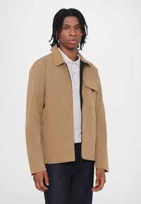 Braune, leichte Jacke mit strukturiertem Design, Taschen auf der Vorderseite und schwarzem Reißverschluss. Getragen über einem hellgrauen Poloshirt und dunklen Jeans.