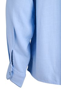 Gaiši zils garo piedurknu krekla uzmava ar pogu un sānu spraugu, kas parāda auduma tekstūru un šuvju detaļas.