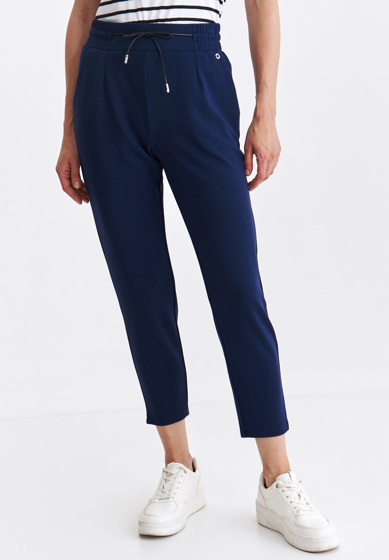 Osoba ubrana w granatowe, krótke spodnie z elastycznym paskiem i sznurkiem, w połączeniu z białymi sneakersami i pasiastą koszulą.