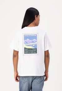 White short-sleeve t-shirt featuring a colorful printed design resembling a postage stamp, depicting mountains and water with text.