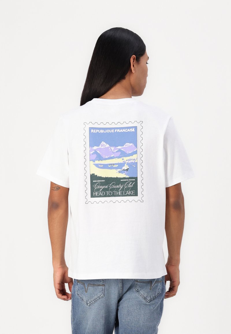 White short-sleeve t-shirt featuring a colorful printed design resembling a postage stamp, depicting mountains and water with text.