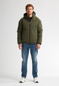 Grüne gepolsterte Jacke mit Reißverschluss und verstellbarem Kapuzen, getragen über einem schwarzen Oberteil und blauen Jeans, mit elastischen Bündchen und lockerer Passform.