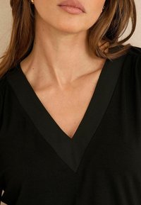 Cămașă neagră cu decolteu în V, realizată dintr-un material neted, cu un tiv distinct în jurul gâtului, subliniind linii curate și un design simplu.