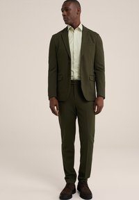 Traje verde oliva con blazer de un solo botón, pantalones a juego y una camisa de botones verde claro. Complementado con zapatos marrones.