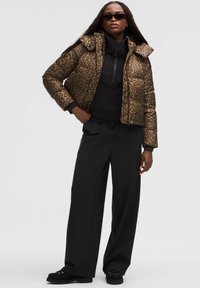 Přesně střižená pufovaná bunda s leopardím potiskem a vysokým límcem, spárovaná s černou zipovou mikinou a volnými černými kalhotami. Model má na sobě černé sluneční brýle.