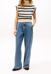 Beżowy sweter na drutach z granatowymi paskami i wysokim kołnierzem, zestawiony z szerokimi jasnoniebieskimi jeansami. Modelka ma ręce w kieszeniach; czarne buty.