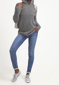 Grijze gebreide hoodie met een hoge hals, gecombineerd met lichtblauwe spijkerbroek. Grijze sportschoenen en een zilveren rugzak maken de look compleet.