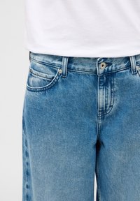 Gaiši zilas džinsa bikses ar izbalējušu apdari, priekšā ar pogu aizdari, standarta kabatām un nelielām nodiluma zīmēm šuvēs.