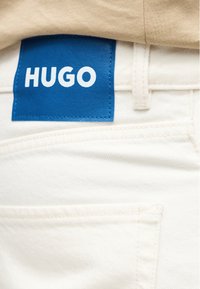 Κοντινό πλάνο σε λευκά τζιν με μπλε δερμάτινη ετικέτα ραμμένη πάνω από την πίσω τσέπη με το όνομα της μάρκας "HUGO" σε λευκά γράμματα.