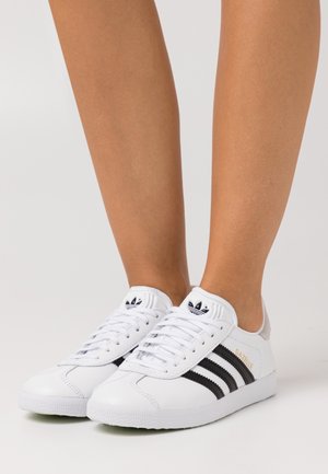 Baskets blanches Adidas Gazelle avec des bandes noires portées aux pieds, présentées sur un fond clair uni.