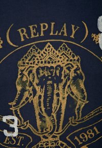 Tmavě modré tričko s potiskem zlatého slona, detailní korunou a textem; zahrnuje bílé číslo "3" a rok založení "1981."