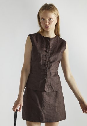 Jeune femme aux cheveux blonds et raides porte un haut sans manches marron foncé boutonné et une jupe assortie, tenant un sac à main noir orné de perles.
