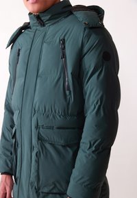 Parka verde imbottita con colletto alto, cappuccio regolabile, tasche anteriori con zip e tasche sul petto con zip verticale; texture liscia.
