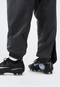 Μαύρα ποδοσφαιρικά παπούτσια με μπλε λεπτομέρειες και το κείμενο «AIR ZOOM», φορεμένα με μαύρο παντελόνι που έχει φερμουάρ στον αστράγαλο πάνω από λευκές κάλτσες.