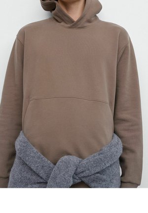 Persona con sudadera con capucha marrón y un suéter gris de punto atado a la cintura, sobre un fondo liso.