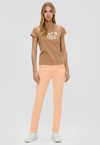 Bruine katoenen t-shirt met korte mouwen en witte bloemgrafiek; perzikkleurige slim-fit broek; witte sneakers; neutrale achtergrond.