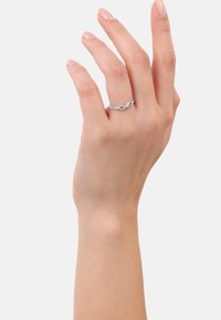 Una mano con pelle chiara mostra un anello d'argento con un design infinito, posizionato su uno sfondo bianco uniforme.