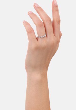 Una mano con pelle chiara mostra un anello d'argento con un design infinito, posizionato su uno sfondo bianco uniforme.